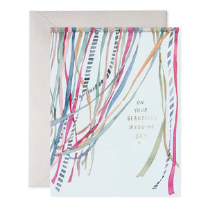 The WEDDING RIBBONS GREETING CARD by E.FRANCES PAPER features vibrant ribbons resembling a watercolor painting on a soft background. Embellished with gold writing, it beautifully states "On your beautiful wedding day." Accompanied by a partially visible envelope, this card completes the delightful wedding greeting experience.