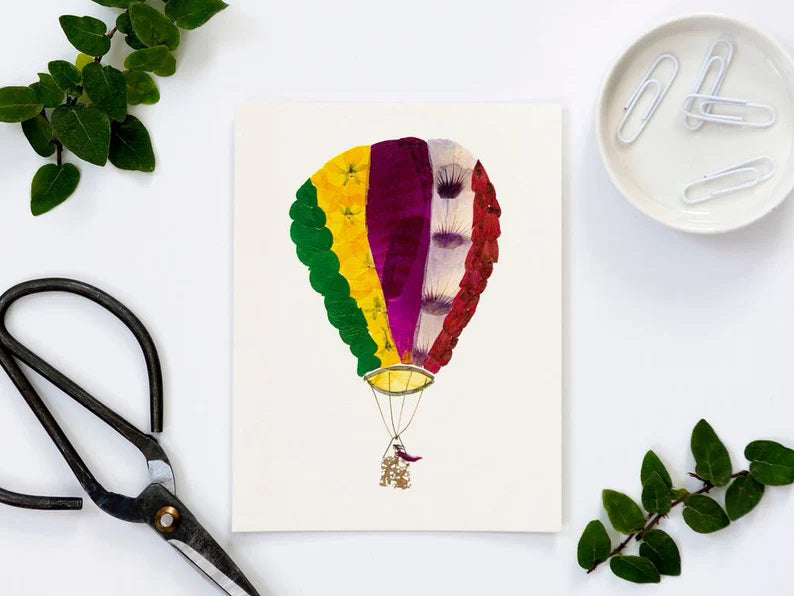 Illustration of a vibrant botanical hot air balloon featuring patchwork panels in shades of green, yellow, purple, and red. The balloon is connected to a small basket with rope against a plain white background, ideal for digital printing on creamy card stock. Featured in the PETAL PEOPLE PRESS - SKY'S THE LIMIT GREETING CARD by Petal People Press.