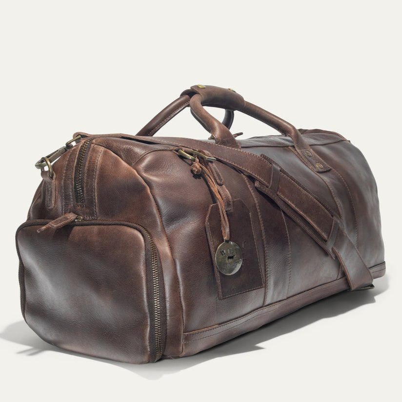 A brown leather duffel bag with sturdy handles, a shoulder strap, brass hardware, and a round metal tag, photographed on a light background.