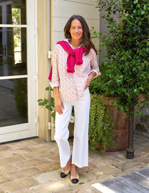 A woman stands outside near a doorway, smiling, her long hair cascading down. She wears FRANK & EILEEN's EILEEN Relaxed Button-Up in Tiny Pink Roses with white pants. Leafy potted plants frame the scene, and the shirt's bust-flattering buttons enhance the natural and peaceful ambiance.