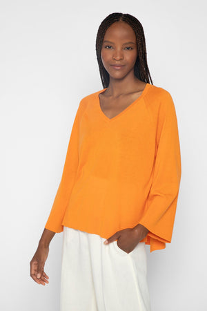 A discerning dresser with long braided hair wears the KINROSS CASHMERE EASY HI LOW VEE NECK SWEATER in orange and white pants. They stand against a plain background, smiling softly, with one hand in their pocket and the other by their side.