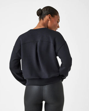 A woman stands confidently against a plain white background, wearing the SPANX Aireessentials Crew Neck Pullover and matching sweatpants made from luxurious comfort spacer fabric. She has her hair pulled back and her left hand is raised slightly. She smiles gently with her lips closed.