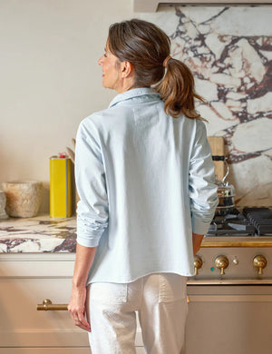 A person stands in a kitchen, hands in pockets, wearing the FRANK & EILEEN PATRICK POPOVER HENLEY IN ICE and white pants, with a stove and kettle behind them. The marbled backsplash displays greenery and a decorative dish on the countertop.