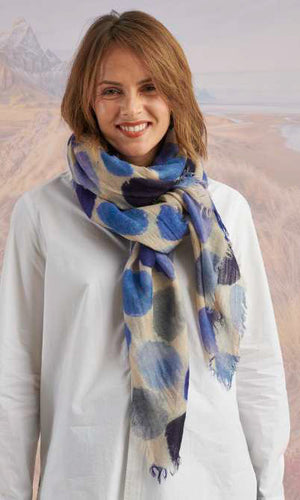 A woman with blonde hair displays the STORIATIPIC - BETTY WOOL SCARF, a large rectangular wool accessory with earth-toned polka dots, set against a scenic mountain landscape.