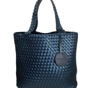 The WOVEN REVERSIBLE TOTE BAG by LINES OF DENMARK is a white tote featuring two shoulder straps and a circular faux-leather tag on one strap. This shopper showcases a textured, interlaced pattern and reversible design, providing stylish and elegant dual styling options. The image background is plain white.