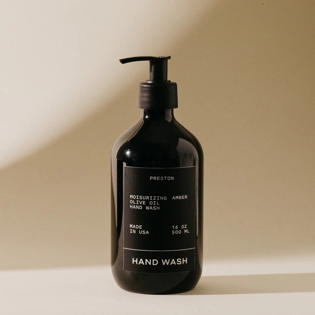 A black pump bottle with a label that reads &quot;PRESTON - HAND WASH 16OZ Moisturizing Olive Oil Hand Wash Amber, Sulfate-Free, Made in USA.&quot; The bottle is standing against a neutral background.