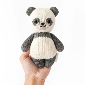 Introducing the CUDDLE + KIND - HANDMADE BABY PANDA from CUDDLE & KIND, a charming, hand-knit plush toy featuring gray ears, arms, and legs, and a white body accented with gray around the eyes and limbs. The panda's smiling face and black eyes add to its adorable appeal. This lovely toy is perfect for baby shower gifts, crafted with love to support fair trade income initiatives.