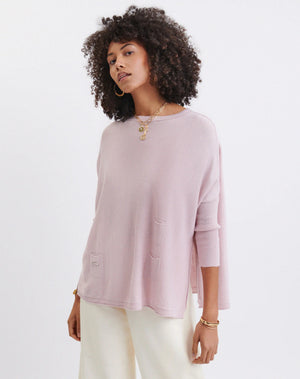 Wearing the MERSEA - CATALINA CREWNECK SWEATER in versatile light pink, a curly-haired individual stands against a plain background. Styled with light-colored pants, hoop earrings, and layered necklaces, they embody the charm of a year-round hero.
