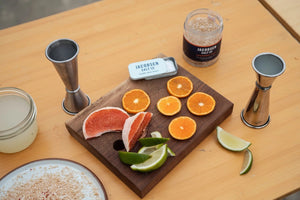 A person is preparing a cocktail at an outdoor setup on a beach. They are pouring Jacobsen Salt Co.'s Jacobsen Salt - Infused Habanero Salt onto a plate from a jar. The setup includes jars of liquid, a small tray with fruit slices, and a scenic backdrop of trees and sand.