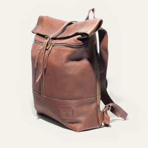 The WILL LEATHER GOODS - ROLL TOP BACKPACK by WILL LEATHER GOODS is open, showing a camera, a red-and-black plaid lining, an Andrzej Sapkowski book, and an old yellowed map inside.