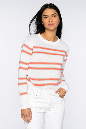 A person with long brown hair stands against a plain white background, wearing a cozy KINROSS CASHMERE - LINEN STRIPE CREW PULLOVER from KINROSS, featuring horizontal blue and gray stripes. They paired it with white pants. Their right hand is in their pocket while their left arm rests by their side.