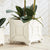 A CHINOISERIE CACHEPOT - LARGE by NAPA HOME AND GARDEN, a square, white ceramic planter featuring an elegant bamboo design, sits on a light wooden surface. The planter holds a green plant with large, glossy leaves. Another similar cachepot is partially visible on the left side of the image, enhancing the home decor ambiance.