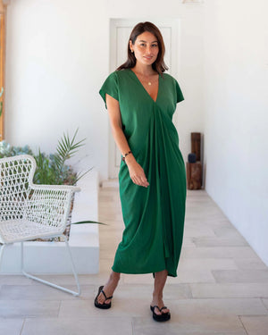 A woman stands in a well-lit hallway, wearing the vibrant MERSEA - IBIZA KAFTAN DRESS, accessorized with a necklace and black sandals. The lightweight green dress is complemented by a white chair and greenery in the background, creating a resort-worthy, modern atmosphere.