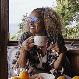 A curly-haired person with glasses from the CADDIS Root Cause Analysis Readers collection sits by a window, holding a mug and smiling. Their aviator-shaped frames catch the light. On the table beside them are a bowl of oranges and a slice of cake. The background shows lush green foliage with a glimpse of the ocean gleaming in the distance.