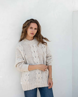 The image displays a detailed view of a person dressed in the MERSEA - TOPANGA CABLE SWEATER by MER SEA, showcasing its chunky knit cream-colored texture with large buttons on the sides, paired with blue jeans. The emphasis is on the sweater's intricate cable design and fine details, with the person's hand partially visible at their side.