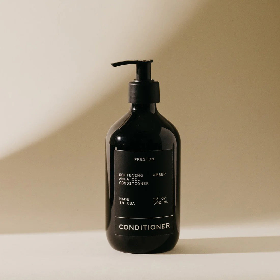 A dark bottle of PRESTON - CONDITIONER with a pump top, labeled "Preston Softening and Shine Conditioner." The label also includes text stating "Made in USA," "16 oz / 500 ml," and "cruelty free." Infused with Indian Amla Oil, this nourishing conditioner is set against a neutral background with a shadow cast on the right.