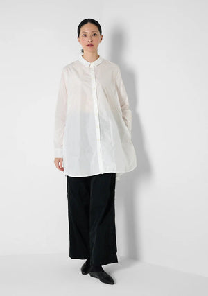 A person with curly hair wears the KATHARINA HOVMAN oversized taffeta blouse, featuring mother-of-pearl buttons, along with black pants. They stand in a minimalist setting with hands in pockets, gazing at the camera.