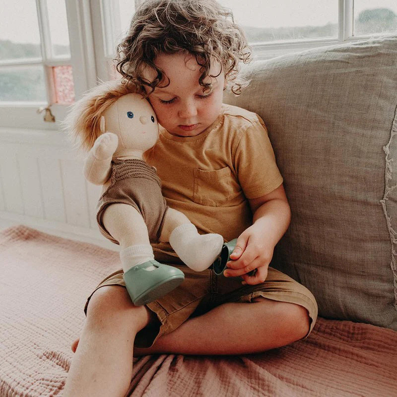 A close-up of a baby wearing OLLI ELLA USA DINKUM DOLL SHOES in Mallow Pink and white socks. The baby is seated on a soft, pale pink textured blanket, dressed in light brown clothing. Only the lower half of the baby&#39;s body is visible.