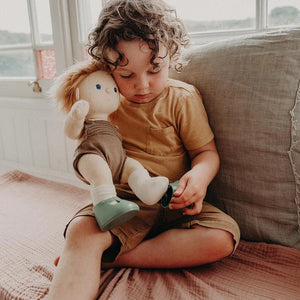 A close-up of a baby wearing OLLI ELLA USA DINKUM DOLL SHOES in Mallow Pink and white socks. The baby is seated on a soft, pale pink textured blanket, dressed in light brown clothing. Only the lower half of the baby's body is visible.