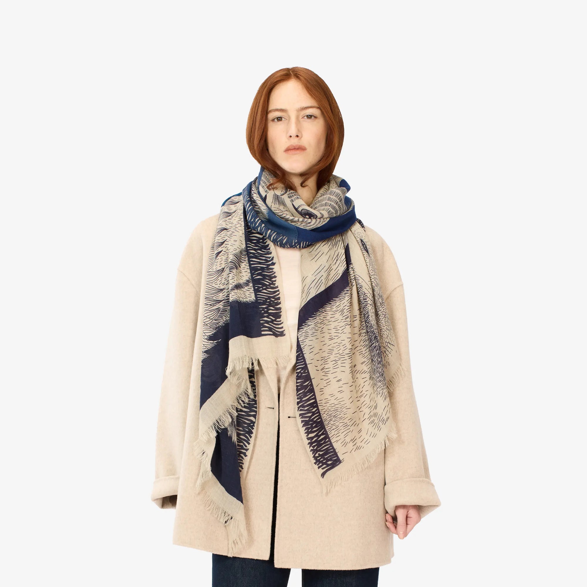 A person with shoulder-length red hair is wearing a light beige coat over a light-colored top, dark pants, and the INOUI EDITIONS - FRERES SCARF by INOUI, which features a detailed face design and is perfect for winter warmth. The background is plain white.