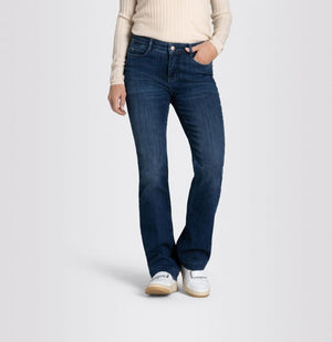 A person wearing a long-sleeve, light-colored ribbed top and MAC's DREAM BOOT AUTHENTIC DENIM boot cut jeans in dark blue with a slight flare at the bottom. They are also wearing white sneakers and have one hand in their pocket, standing against a plain, light gray background.