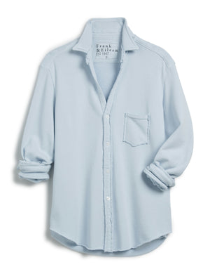 A light blue long-sleeve button-up shirt with a collar and single chest pocket is laid flat, its sleeves casually rolled up to highlight the relaxed fit. The inner label reads "FRANK AND EILEEN." Product: EILEEN SWEATSHIRT BUTTON UP IN ICE by FRANK & EILEEN.