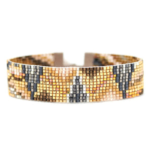 The JULIE ROFMAN - ORO BRACELET by JULIE ROFMAN JEWELRY is a woven piece adorned with a geometric pattern in shades of brown, beige, and black. It features handwoven Delica beads and includes a suede tie closure for adjustable sizing. The bracelet is displayed on a plain white background.