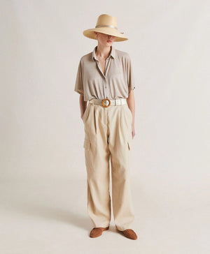 A woman with platinum blonde hair stands with a neutral expression, wearing the MOMONI - BROOKLYN LUREX JERSEY BLOUSE and colorful beaded necklaces. She has her hands in the pockets of her striped trousers with oversize lines. The background is plain and softly lit.