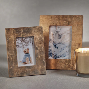 Two GOLD LEAF PHOTO FRAME - 5x7 by ZODAX, perfect for displaying photographs, are placed on a gray surface. One frame features an image of a dog in a blue sweater against a snowy background, while the other displays snow-covered evergreen branches. A lit gold candle nearby enhances the scene with its warm glow.