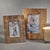 Two GOLD LEAF PHOTO FRAME - 5x7 by ZODAX, perfect for displaying photographs, are placed on a gray surface. One frame features an image of a dog in a blue sweater against a snowy background, while the other displays snow-covered evergreen branches. A lit gold candle nearby enhances the scene with its warm glow.