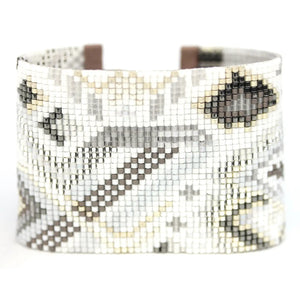 The JULIE ROFMAN - QUARTZ CUFF by JULIE ROFMAN JEWELRY is a wide bracelet showcasing a handwoven, beaded geometric pattern in neutral tones. It features two beige leather straps with silver buckles and an open rectangular center for a distinctive touch.