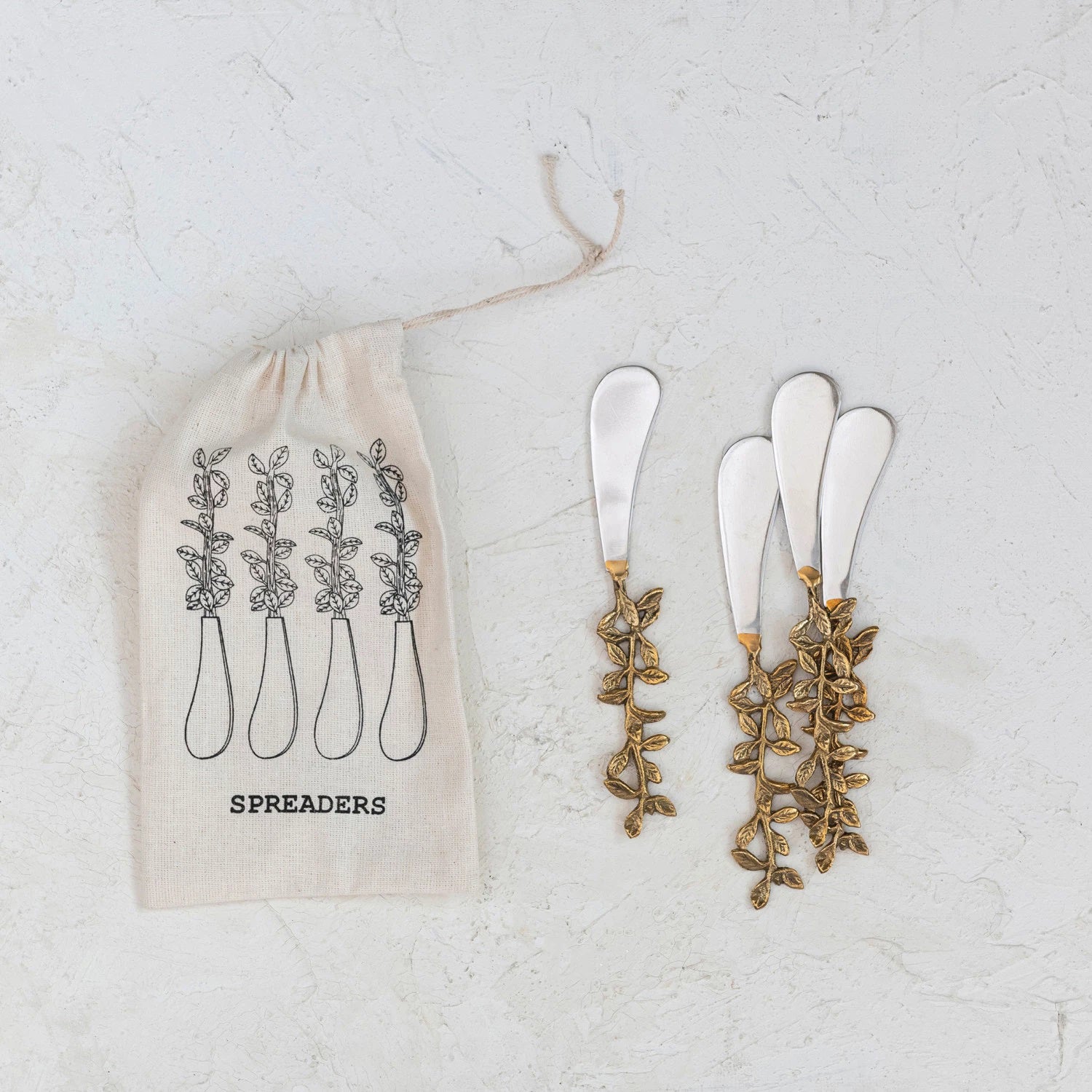 A set of four STAINLESS STEEL AND BRASS LEAVE HANDLE CANAPE KNIVES from CREATIVE COOP is elegantly displayed on a light textured surface. Nearby, there's a cloth pouch labeled "Spreaders" featuring an illustration of the utensils.