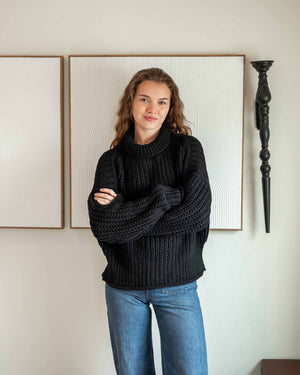 A woman with curly hair stands against a white wall, wearing a chunky mustard-colored MERSEA - BEACHSIDE TURTLENECK and blue jeans. She holds the hem of her turtleneck with one hand and has a relaxed expression.