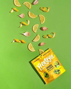 A yellow packet of FREESTYLE - LEMON GARLIC GREEN OLIVES from FREESTYLE SNACKS, featuring green olives flavored with extra virgin olive oil. The packaging highlights the "all-natural" ingredients and notes that the olives are "grown in Greece." Weighing 4 oz (113g), the pack showcases images of green olives alongside phrases such as "pitted & unpasteurized" and "healthy snacking on the go.