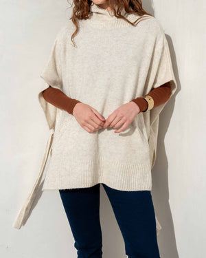 A person standing against a plain wall is dressed in the MERSEA Cape Poncho Sweater, made from a beige cotton knit blend, layered over a brown long-sleeve shirt and paired with dark blue pants. With one hand resting over the other, the poncho’s loose fit and ribbed hem provide both comfort and style.