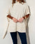 A person standing against a plain wall is dressed in the MERSEA Cape Poncho Sweater, made from a beige cotton knit blend, layered over a brown long-sleeve shirt and paired with dark blue pants. With one hand resting over the other, the poncho’s loose fit and ribbed hem provide both comfort and style.