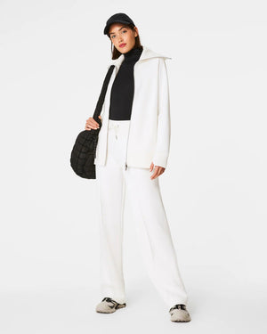 Someone with shoulder-length hair wears a white SPANX AirEssentials Luxe Full Zip Tunic Jacket with matching pants, hands in the tunic's pockets. The plain white backdrop highlights the minimalist loungewear aesthetic.