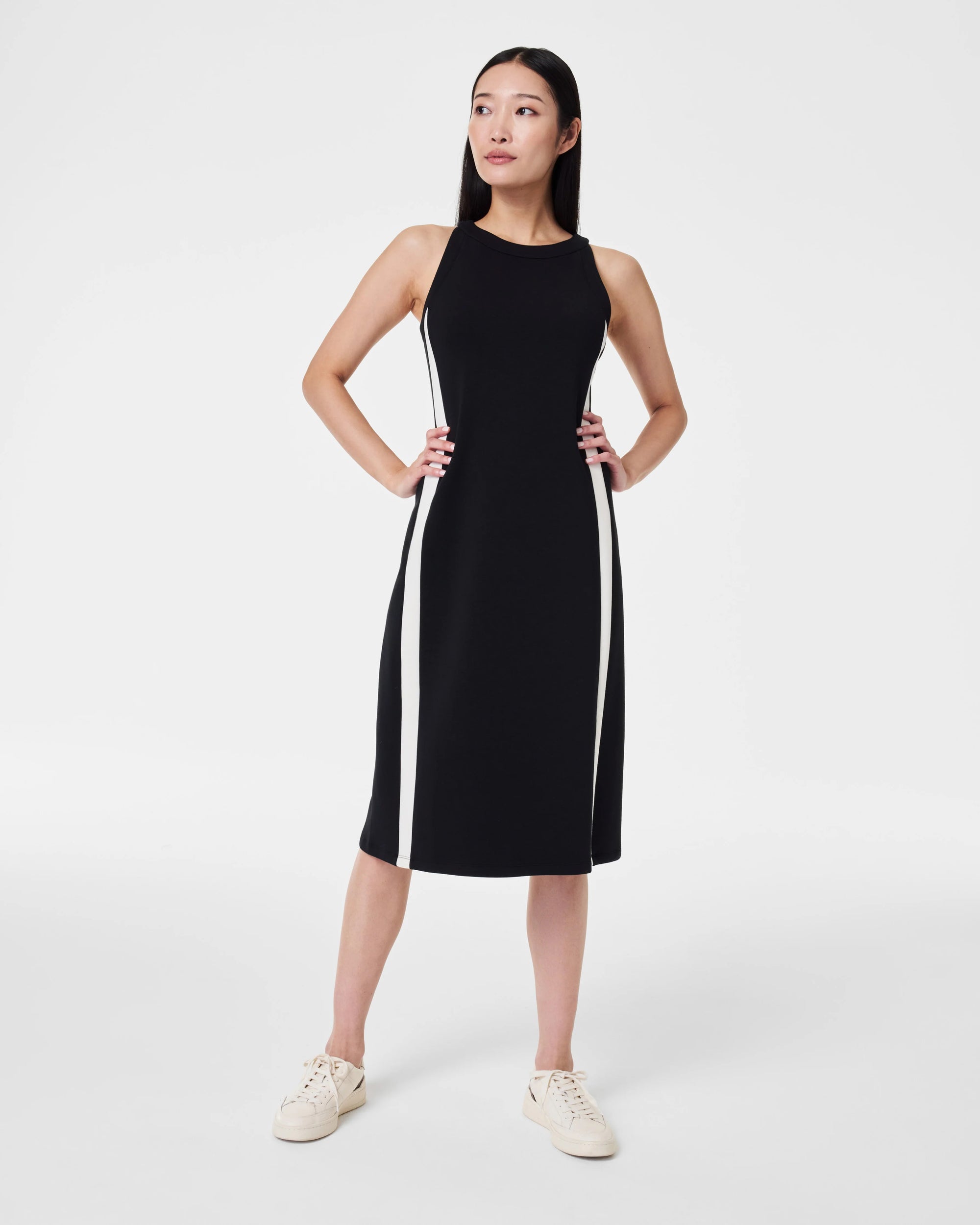 A person with long dark hair is standing against a plain white background, facing away from the camera. They are wearing the SPANX - AIRESSENTIALS SIDE STRIPE MIDI DRESS, a sleeveless black knee-length piece crafted from light-as-air fabric. The dress is stylishly paired with white sneakers, offering an elegant blend of comfort and style.