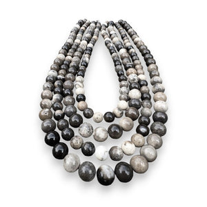 The ANN LIGHTFOOT - FOUR STRAND SHEEP SKIN JASPER NECKLACE by ANN LIGHTFOOT features a collection of multistrand necklaces composed of round, polished stone beads in varying shades of black, gray, brown, and white. These are paired with adjustable nylon ties for a custom fit. When laid out on a weathered wooden surface, the beads exhibit natural patterns and textures that evoke the beauty of sheep skin jasper.