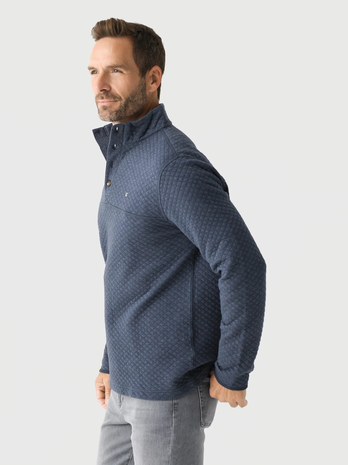 A man with short brown hair and a beard smiles while wearing the CRIQUET - QUILTED PULLOVER IN HEATHER NAVY over a blue plaid shirt and light gray jeans, standing against a plain white background.