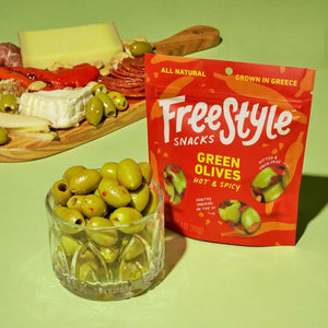 A vibrant orange snack bag labeled "FREESTYLE SNACKS" contains "FREESTYLE - GREEN OLIVES- HOT & SPICY," featuring spicy Calabrian chili. The 4 oz (113g) bag showcases images of green olives with red seasoning and highlights that the product is all-natural and grown in Greece.