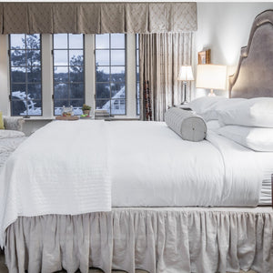 A cozy bedroom featuring a large, neatly made bed adorned with white linens, pillows, and a grey bolster pillow atop the luxurious Kingsdown Fearrington Lifestyle Bedding Collection Queen Pillowtop Mattress with individually wrapped coils. Large windows with curtains in neutral tones offer a view of an outdoor area. A bedside lamp and a small potted plant sit on the nightstand.