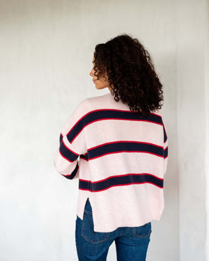 A person with curly hair stands against a light wall, wearing the ultra-soft MERSEA - KEEPER CREWNECK SWEATER in light pink with black and red stripes, paired with blue jeans. The individual faces away, looking to the left.