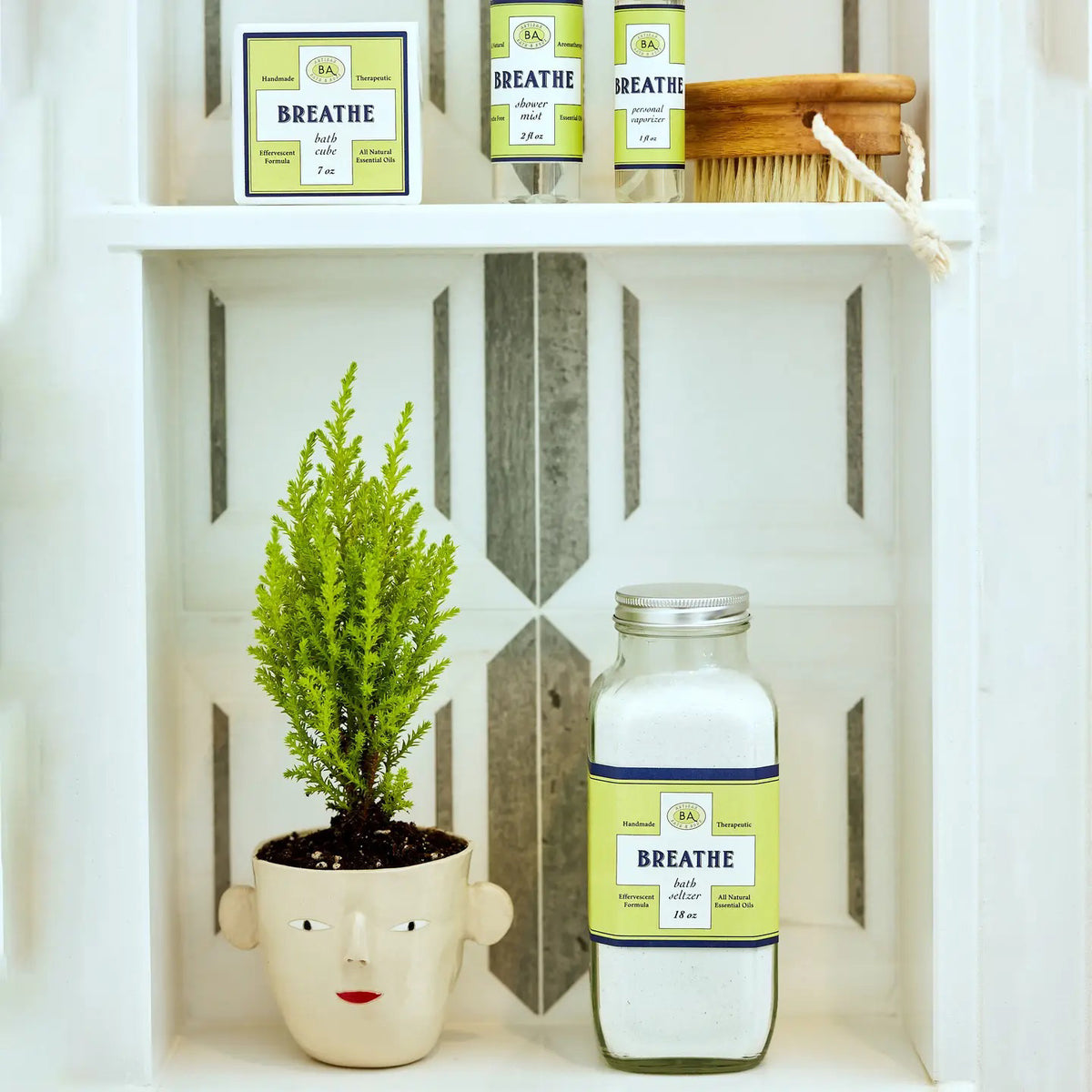 A small green plant in a face-shaped pot sits next to a large jar labeled &quot;BAUDELAIRE - BREATHE SHOWER MIST.&quot; Above, smaller bottles of eucalyptus essential oil and a wooden brush rest on a white shelf with a gray geometric background, creating an inviting space for your aromatherapy steam room.