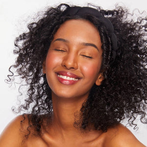 A person with long blonde hair is smiling and looking to the side. They are wearing the KITSCH PUFFY HEADBAND, a black padded accessory crafted from recycled polyester. The plain white background accentuates their cheerful expression and the eco-friendly headband.