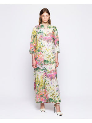 A person stands with their back turned, wearing the MIRTO 1956's Linen Brush Stroke Print Dress featuring floral watercolor patterns in pink, green, and yellow. They have long brown hair and are donning black high heels against a plain white background.