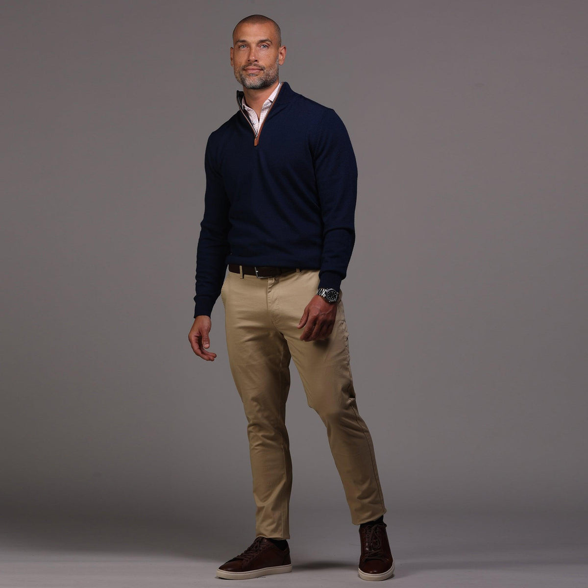 A man with short hair and a beard stands against a gray background wearing the COLLARS AND CO Navy with Brown Suede Trim Cashmere Touch Australian Merino Quarter Zip Sweater over a white shirt, khaki pants, and brown high-top sneakers, one hand in his pocket.