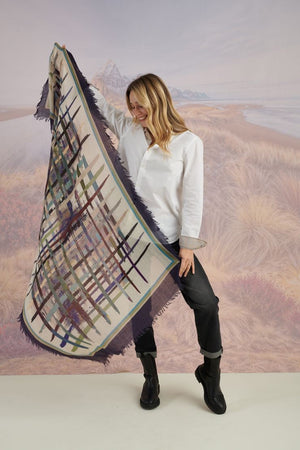 A woman in a blue shirt and jeans stands holding the STORIATIPIC - JULIE WOOL SCARF, a luxury plaid accessory in earthy tones. Behind her, mountains, beach, and grassy dunes create a scenic backdrop.