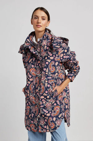 A woman posing confidently with her hands in the pockets of a long, dark floral-patterned ADROITE ATELIER - RYAN RAINCOAT, which features intricate red, orange, and white paisley designs and a full zip closure. The coat complements her straight dark hair and light blue jeans against a plain light grey background.