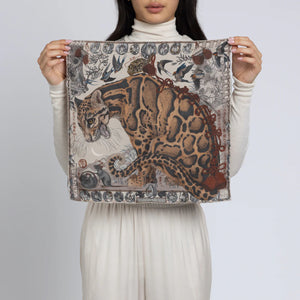 Dressed in cream, a person displays the SABINA SAVAGE - THE LUNAR LEOPARD SILK TWILL SCARF 42CM from SABINA SAVAGE ENGLAND, showcasing an elaborate design of a stylized clouded leopard amid intricate forest elements and animals, evocative of Edo-period Japanese art, all set against a neutral background.